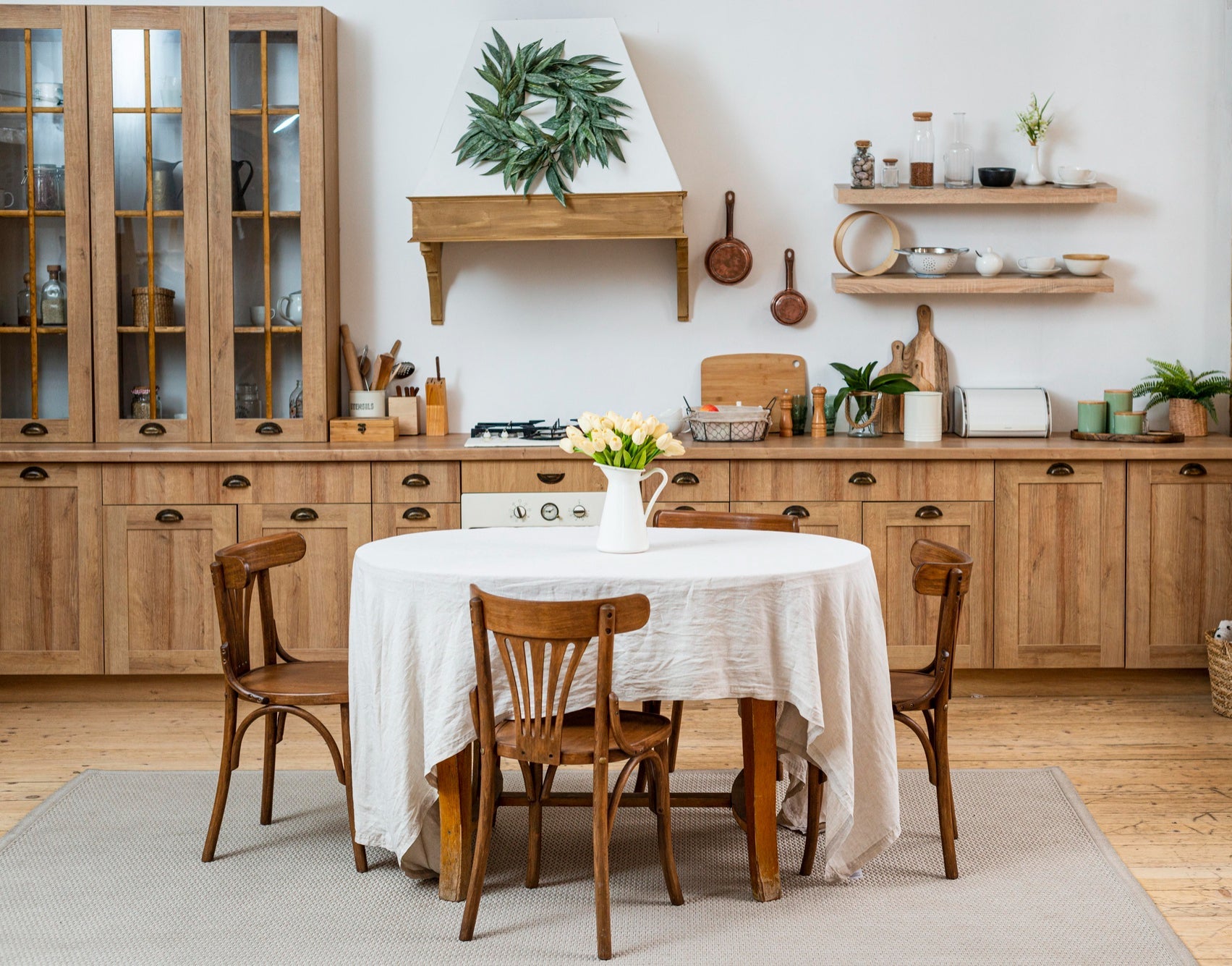 Bright kitchen with wooden cabinets and shelves, featuring a round table set with a white tablecloth and a vase of tulips.