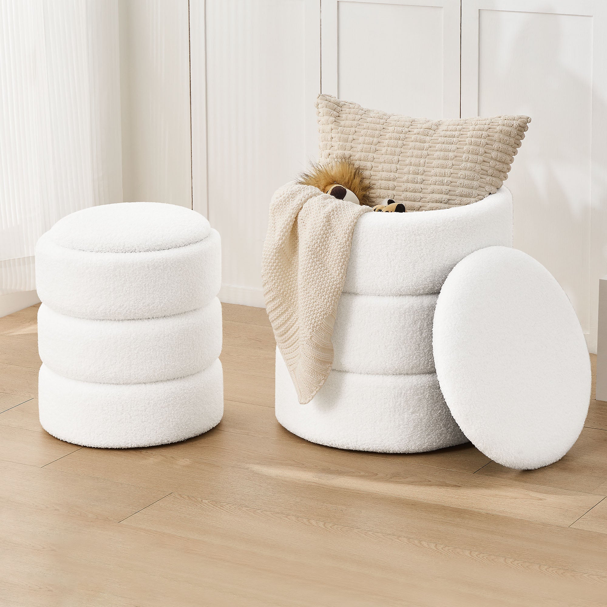 Two white, fluffy-style storage ottomans sit on a wooden floor; one is open with a pillow and blanket inside, the other is a stool.