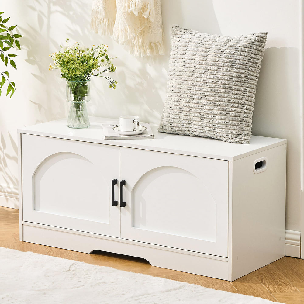 A stylish white storage bench with two black handles, adorned with a vase of flowers, a cushion, and a coffee cup on top.