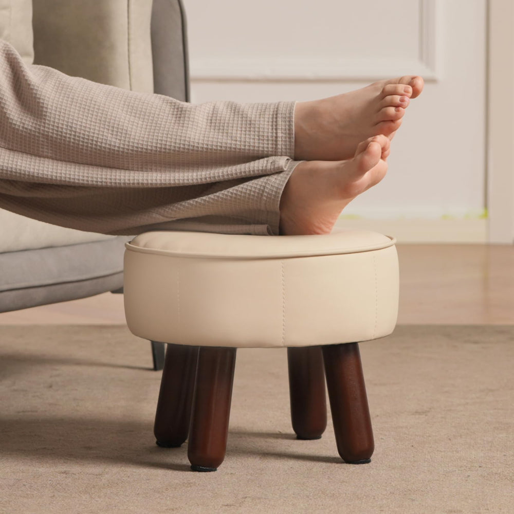 A round beige leather ottoman with a button-tufted top and sturdy wooden brown legs. Ideal for seating or decor.