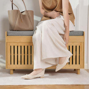 A woman in a beige top and white pants sits on a wooden bench, adjusting her stylish pearl-embellished flat shoes. A beige bag is nearby.