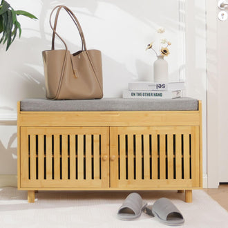 A stylish wooden bench with a gray cushion, a handbag, books, and a vase of flowers, placed near a door and slippers on the floor.