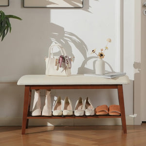 A wooden bench with a cream cushion, holding a handbag and flowers, displays a variety of stylish shoes underneath in a cozy entryway.