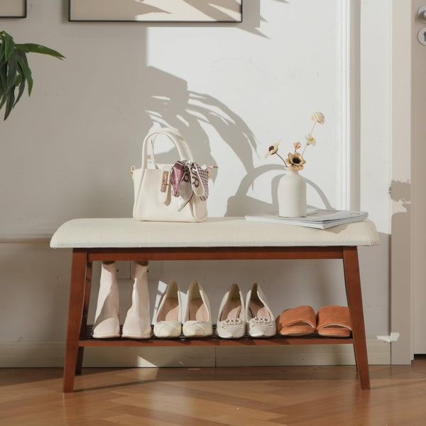A wooden bench with a cream cushion, holding a handbag and flowers, displays a variety of stylish shoes underneath in a cozy entryway.