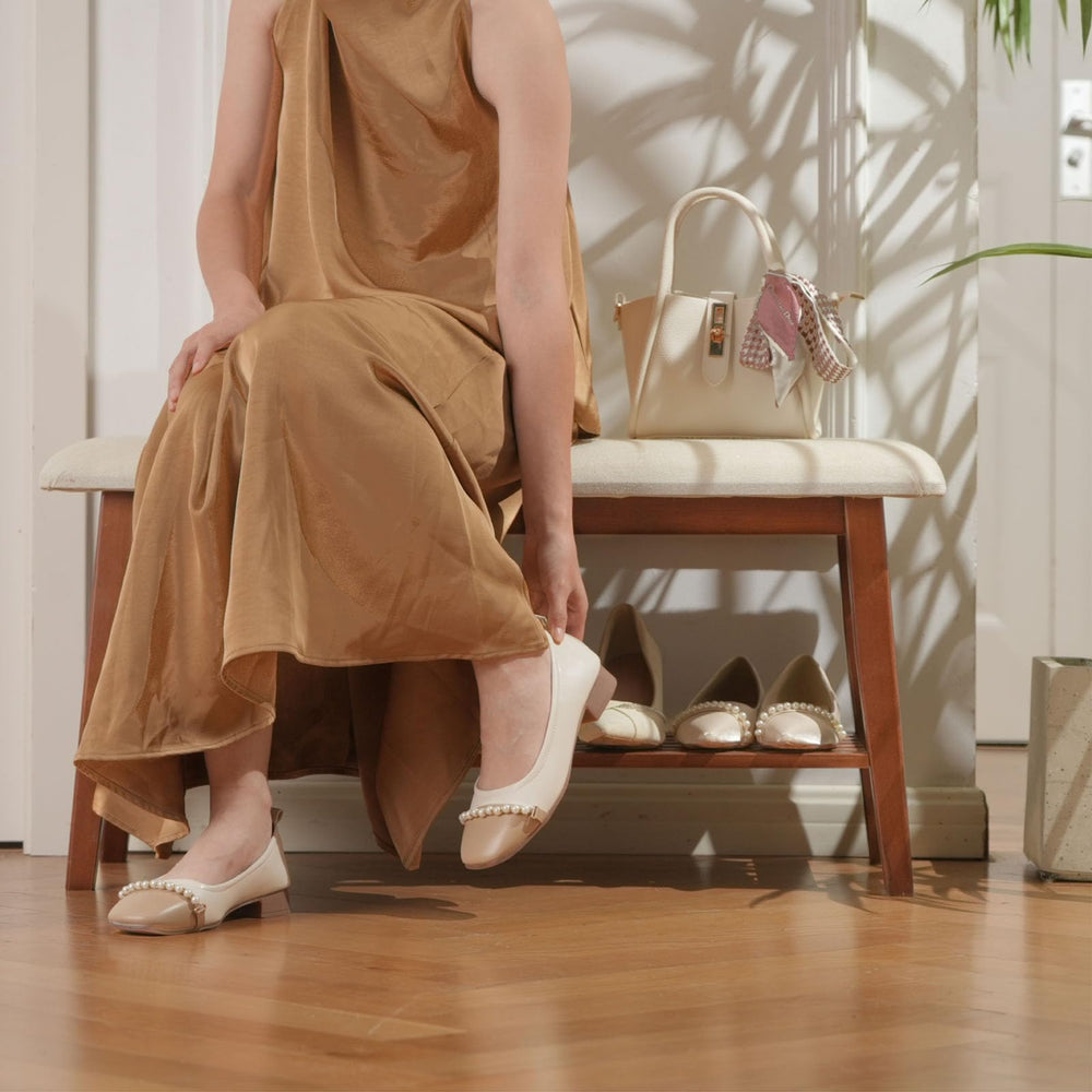 A stylish wooden bench displays elegant shoes and a handbag, with a minimalist vase of dried flowers beside a magazine on top.