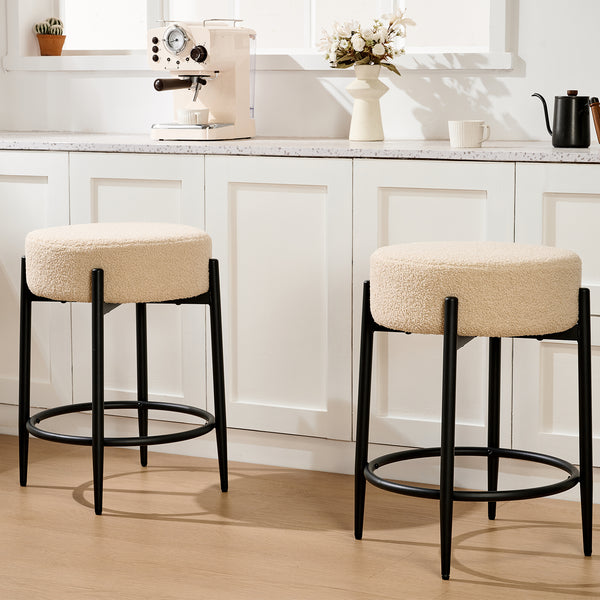 Two beige upholstered bar stools with black legs sit in a modern kitchen, adjacent to a coffee machine and decorative elements.