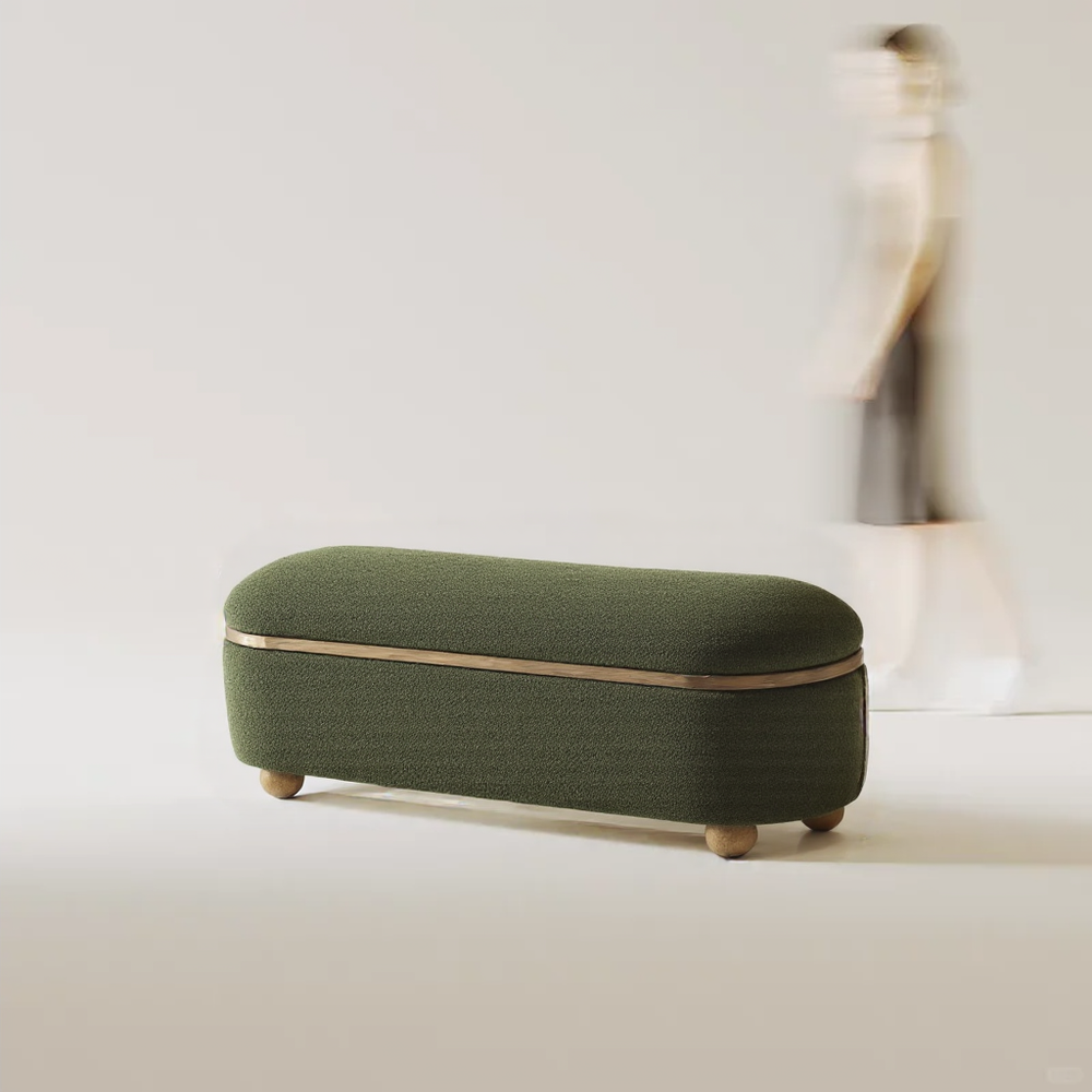 A plush green upholstered storage bench with rounded edges and wooden feet, next to a blurred figure walking in a minimalist setting.
