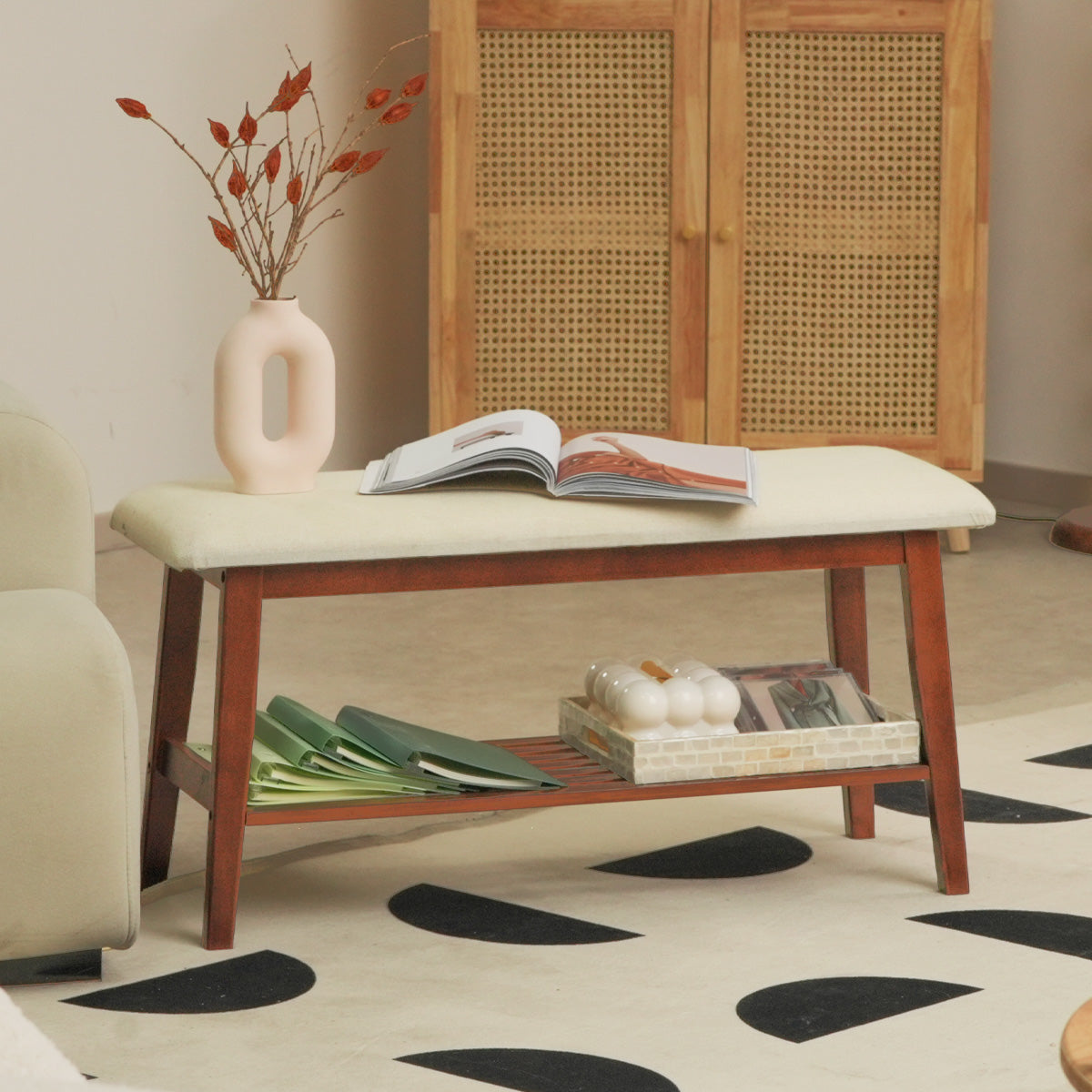 A stylish wooden bench with beige upholstery, a vase of dried branches, and a few books, placed on a patterned rug in a cozy living space.