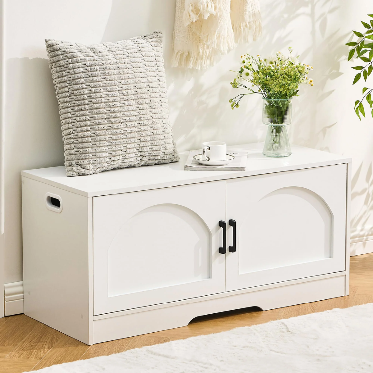 A white storage bench with black handles, topped with a grey textured pillow, a cup, and a vase of flowers, set in a cozy room.