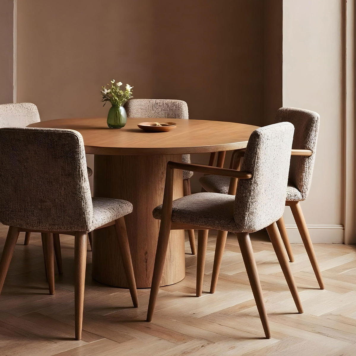 A round wooden dining table with four textured chairs, a vase of flowers, and a wooden bowl, set in a cozy, neutral-toned room.