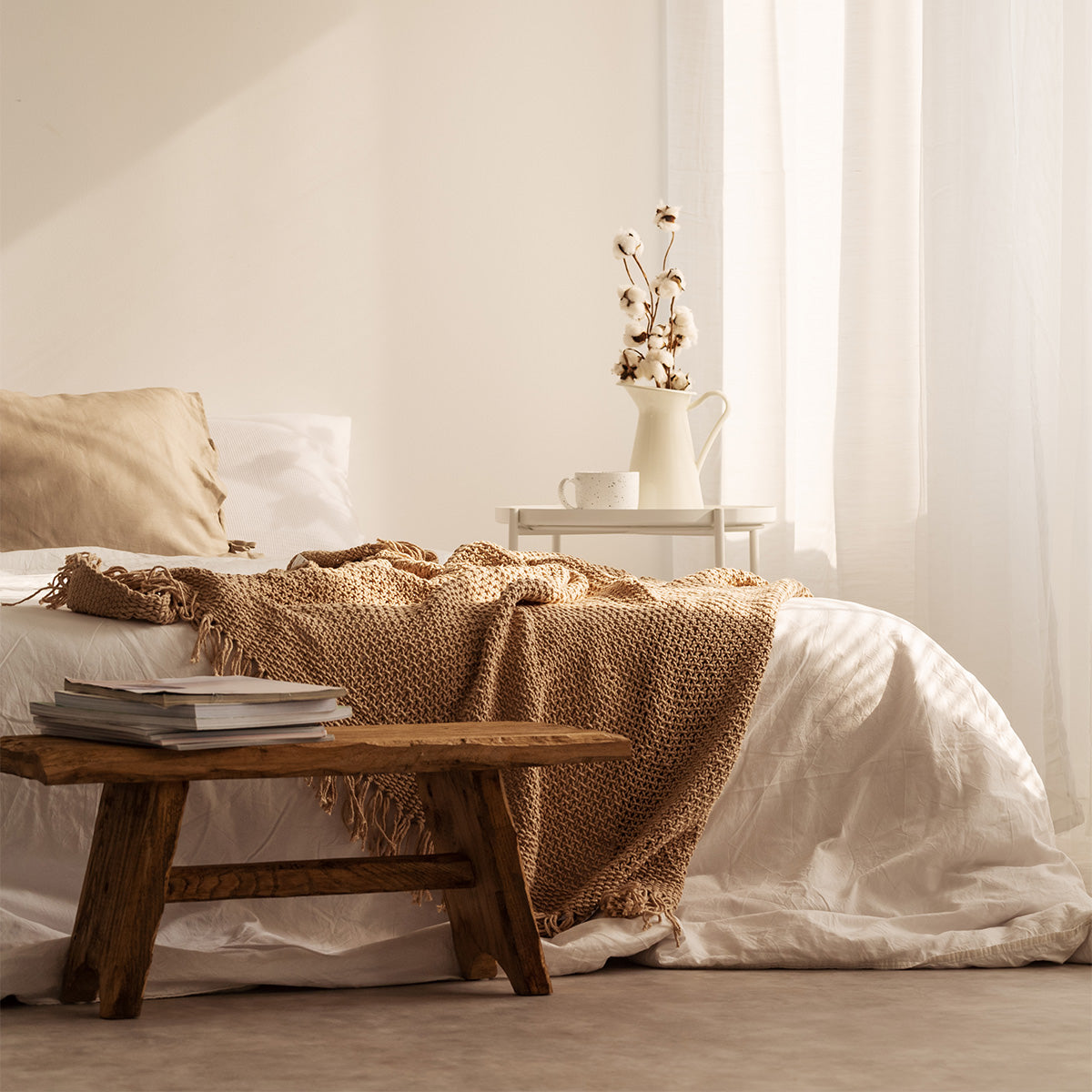 A cozy bedroom scene featuring a white bed with a beige throw, a wooden stool with books, and a vase of cotton flowers. Natural light floods the room.